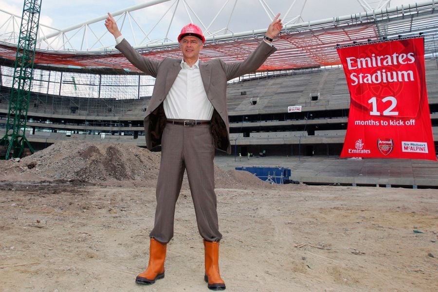 Wenger at Emirates Stadium which is being built. The pitch is bare earth. Wearing a brown suit, hard hat and workboots. His arms are raised