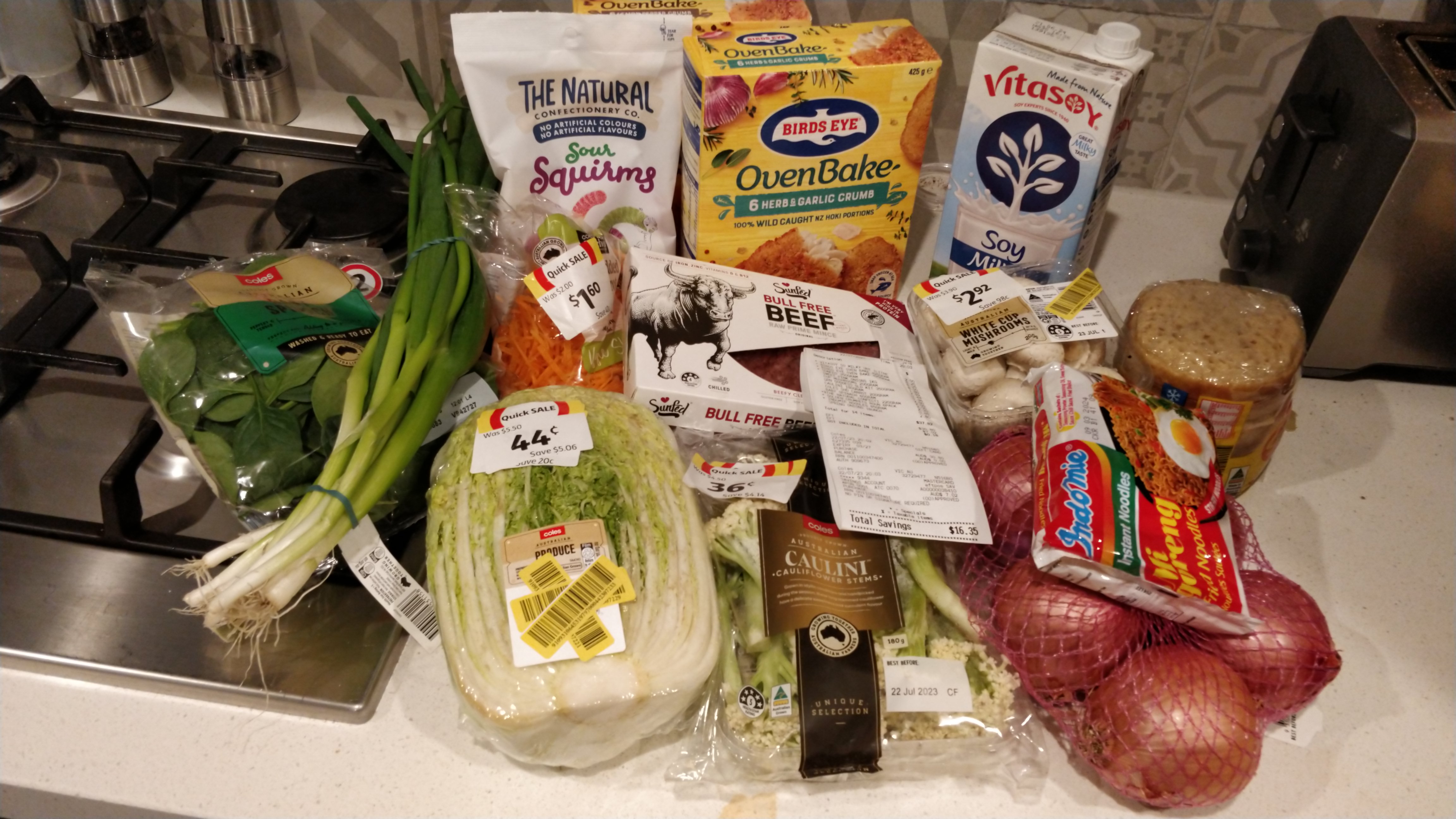 Pile of groceries on kitchen counter - including several marked down veg, two boxes of frozen fish pieces, a bag of sour snakes, a packet of soy milk and a tray of vegan mince