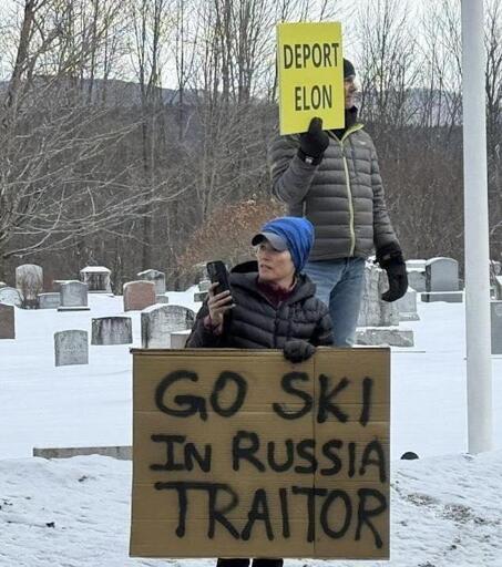 Two protesters stand in a snowy cemetery holding signs. One person in a blue beanie and black puffer jacket holds a large cardboard sign reading "GO SKI IN RUSSIA TRAITOR" in bold black letters. Another protester behind them, wearing a gray puffer jacket and black gloves, holds a yellow sign that says "DEPORT ELON." The setting includes gravestones and bare trees in the background, adding an eerie contrast to the political demonstration.