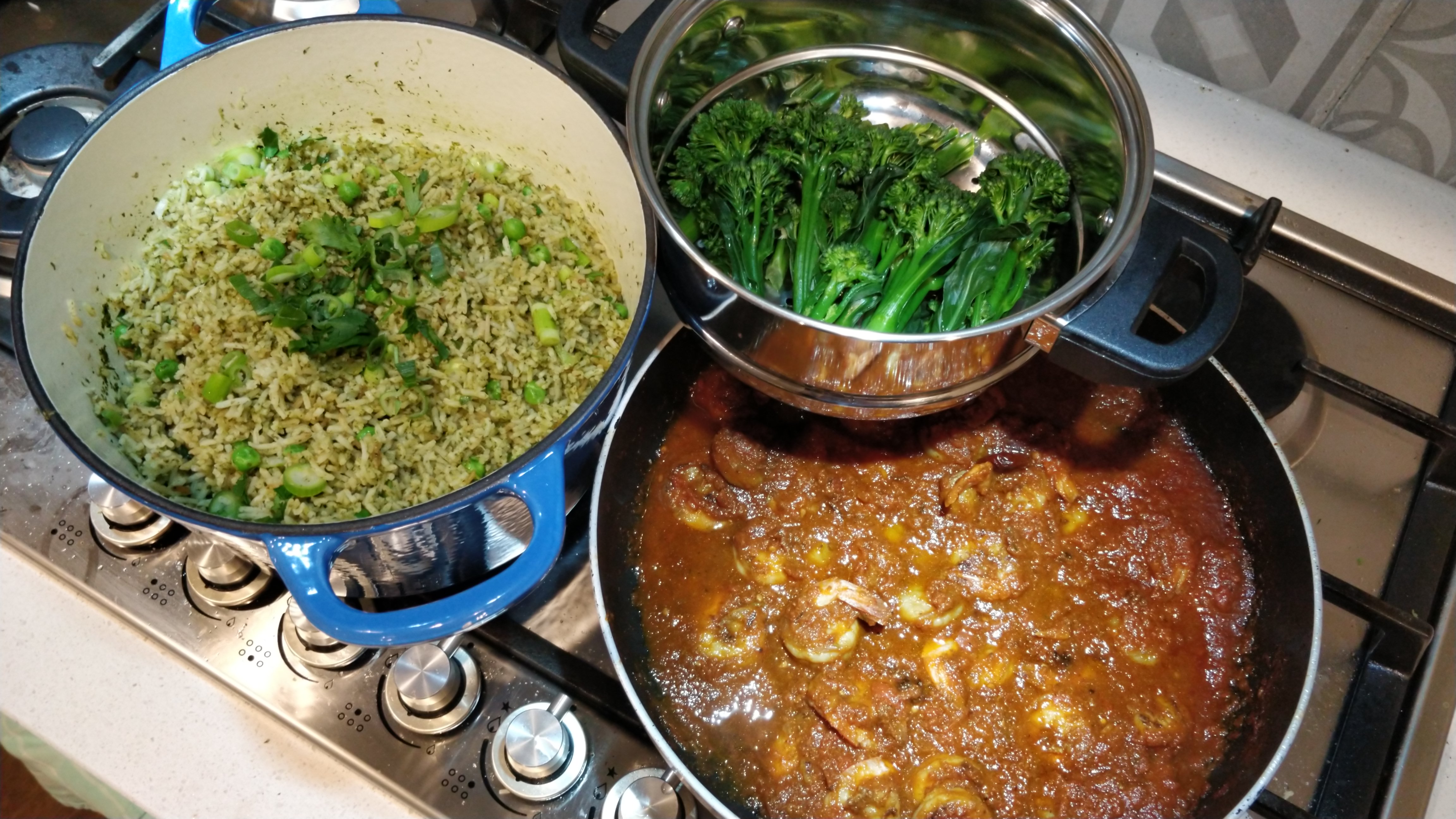 a blue Dutch oven pot with herbed rice, a pan of prawns in a luscious tomato sauce, and a metal strainer with steamed broccolini