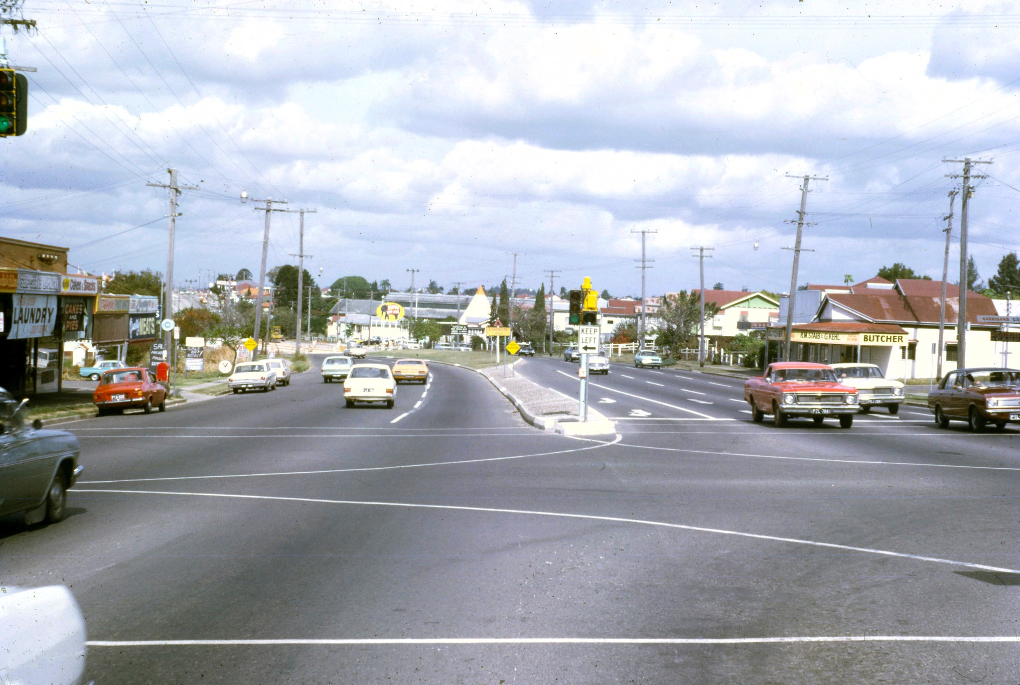 A photo of an intersection on a wide road with old-timey cars driving on the left. In the distance the road curves to the left and follows up a hill.