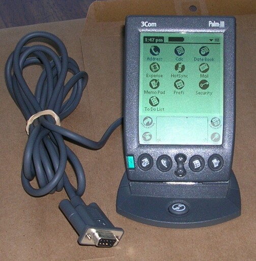 A photo of a Palm III on its docking station. It resembles a large smartphone with a black and white screen, and four large buttons at the bottom.
A serial cable is connected to the docking station.